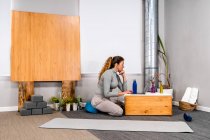Вид сбоку сосредоточенной женщины с длинными волосами в повседневной одежде, работающей на ноутбуке, сидящей на полу за маленьким деревянным столом в студии минимализма йоги — стоковое фото