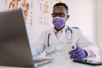 Médecin afro-américain concentré méconnaissable en peignoir médical et masque buvant du café et travaillant sur ordinateur portable dans une clinique moderne — Photo de stock