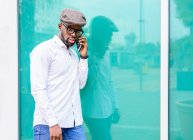 African American male in trendy outfit standing near glass building in city and talking on mobile phone — Stock Photo