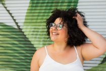 Conteúdo adulto com sobrepeso feminino em óculos tocando cabelo encaracolado contra a parede ornamental durante o dia — Fotografia de Stock