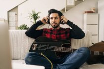 Взрослый музыкант в наушниках играет на бас-гитаре против нетбука на диване в гостиной — стоковое фото