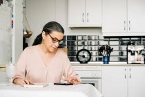 Konzentrierte Frau mittleren Alters in Brille surft Handy, während sie am Tisch mit Notebook in der modernen Küche mit Geschirr zu Hause sitzt — Stockfoto