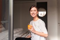 Contenuto giovane donna in abiti casual sorridente e bere succo fresco seduto su un comodo letto vicino alla finestra mentre guarda la fotocamera — Foto stock