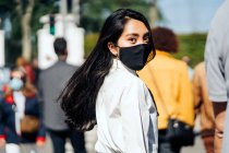 Vista posteriore della femmina asiatica con i capelli volanti che indossano una maschera protettiva che cammina tra la folla e guarda la fotocamera — Foto stock