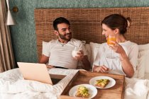 Feliz joven pareja multiétnica en ropa casual desayunando y viendo películas en el portátil mientras está sentado en la cama cómoda durante el fin de semana perezoso en casa - foto de stock
