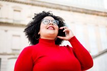 From below of content overweight female in red clothes and eyeglasses talking on cellphone while looking up in city — Stock Photo