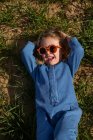 De arriba chica en ropa de moda y gafas de sol de la mano detrás de la cabeza y relajarse en el césped cubierto de hierba - foto de stock