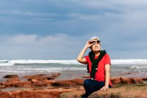 Smiling elderly female tourist in sunglasses sitting on rough boulder while looking up against ocean under cloudy sky — Stock Photo
