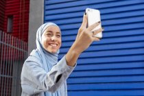 Дружелюбная этническая женщина в платке делает автопортрет на мобильном телефоне на городской улице в солнечном свете — стоковое фото