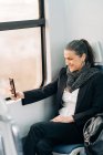 Вид сбоку позитивной женщины средних лет с шарфом, которая фотографируется на мобильный телефон, сидя на пассажирском сиденье у окна в самолете во время поездки — стоковое фото