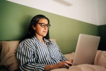 Ethnische Freiberuflerin mittleren Alters mit langen dunklen Haaren in lässiger Kleidung und Brille sitzt im Bett und arbeitet zu Hause ferngesteuert am Laptop — Stockfoto