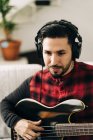 Erwachsener männlicher Musiker mit Kopfhörer spielt Bassgitarre gegen Netbook auf Sofa im Wohnzimmer — Stockfoto