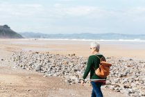 Вид ззаду на невизначену старшу жінку-похідницю, що йде на піщаному березі проти нескінченного моря під час подорожі — стокове фото