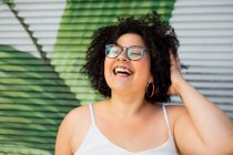 Conteúdo adulto com sobrepeso feminino em óculos tocando cabelo encaracolado contra a parede ornamental durante o dia — Fotografia de Stock