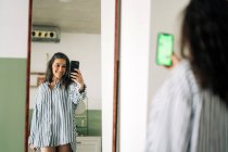 Веселая женщина средних лет в полосатой блузке, отражающейся в зеркале во время съемки автопортрета на смартфоне в комнате дома — стоковое фото