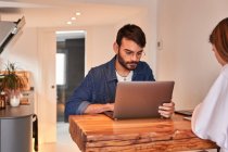 Freelancer masculino hispânico focado jovem trabalhando remotamente em laptop sentado à mesa na cozinha com namorada navegando smartphone — Fotografia de Stock