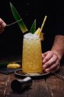 Анонимный бармен, собирающий спиртный коктейль с зелеными листьями и ананасом на подносе возле рюмки за деревянным столом на черном фоне — стоковое фото