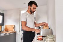 Vista lateral de un joven hombre hispano barbudo con ropa casual haciendo tostadas crujientes mientras prepara el desayuno en casa por la mañana. - foto de stock