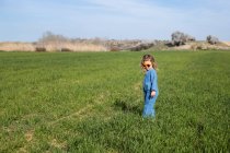 Вид збоку мила щаслива дівчинка в модному одязі і сонцезахисних окулярах стоїть і розслабляється на трав'янистому газоні — стокове фото