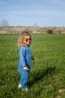Vue latérale de mignonne petite fille heureuse dans des vêtements à la mode et des lunettes de soleil debout et relaxant sur pelouse herbeuse — Photo de stock