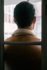 Vista trasera del macho con el torso desnudo de pie cerca de la ventana en casa y mirando hacia otro lado - foto de stock