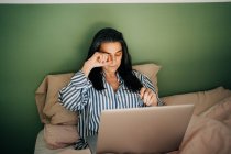 High angle of exhausted middle aged Hispanic female freelancer rubbing eyes while sitting on bed and working remotely on laptop at home — Stock Photo