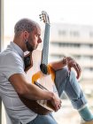 Pensive male with tattooed bald head in casual clothes sitting on windowsill and playing guitar in daytime — Stock Photo