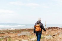 Back view of unrecognizable elderly female backpacker with trekking pole strolling on boulders against stormy ocean under cloudy sky — Stock Photo