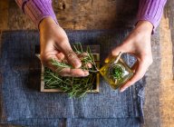 Top view of crop unrecognizable female demonstrating small wooden chest filled with herbs twigs with green leaves near essential oil glass bottles on textile near scissors at table — Stock Photo