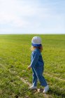 Full body side view of unrecognizable girl in stylish clothes and cap looking away while standing on grass on sunny summer day in field — Stock Photo