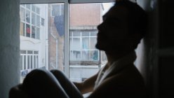 Vista lateral de las rodillas masculinas pensativas abrazando y sentado en el alféizar de la ventana en casa - foto de stock