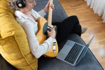 Vue arrière grand angle du guitariste féminin dans les écouteurs jouant de l'instrument de musique sur le canapé avec netbook à la maison — Photo de stock