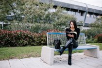 Стильная предпринимательница сидит на скамейке и делает заметки в органайзере во время работы в городском парке — стоковое фото