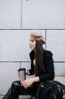Seitenansicht der entzückten stilvollen Unternehmerin im trendigen Outfit und Baskenmütze, die auf der Treppe mit Getränken in der Stadt sitzt und wegschaut — Stockfoto