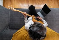 Высокоугольный вид сзади неузнаваемой женщины-гитариста в наушниках, играющей на музыкальном инструменте на диване с нетбуком дома — стоковое фото