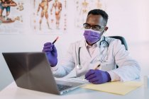 Неузнаваемый серьезный черный ученый в медицинском халате и маске демонстрирует образец крови в пробирке во время видеозвонка на ноутбуке в клинике — стоковое фото