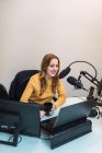 Оптимістична жінка сидить за столом з комп'ютерами і п'є гарячий напій, розмовляючи з мікрофоном під час роботи на сучасній радіостанції — стокове фото
