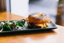 Sanduíche de bagel apetitoso com queijo e frango servido em prato com salada de rúcula na mesa no café — Fotografia de Stock