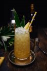 Bicchiere di cocktail alcolico decorato con pezzo di ananas e foglie con paglia di carta su vassoio vicino allo shaker e bottiglia con bicchierino a tavola su sfondo nero — Foto stock