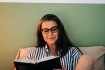 Entspannte reife hispanische Frau in lässiger Kleidung und Brille sitzt im Bett und liest vor dem Schlafen ein interessantes Buch — Stockfoto