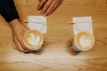 From above of crop unrecognizable person taking takeaway cup of coffee with latte art placed on wooden table in cafe — Stock Photo