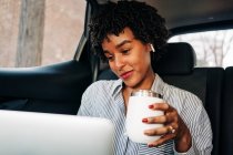 Young content cheerful African American female working with netbook and having mug of coffee to go riding on backseat in car — Stock Photo