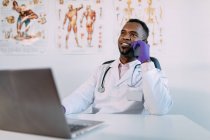 Cheerful young African American male physician in medical apparel and gloves having phone conversation and smiling while sitting at table with laptop and coffee cup during break in clinic — Stock Photo