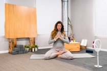Full body of concentrated young female in activewear sitting in Lotus pose with closed eyes and prayer hands doing meditation during yoga session at home near smartphone placed on tripod — Stock Photo
