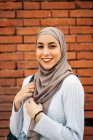 Mulher muçulmana encantadora em lenço de cabeça tradicional e roupas da moda em pé na rua contra a parede de tijolos e olhando para a câmera — Fotografia de Stock