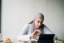 Zufriedene Muslimin im Hijab winkt mit der Hand und spricht im Videochat per Tablet, während sie am Tisch im Café sitzt — Stockfoto