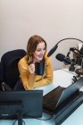Donna ottimista seduta a tavola con i computer e che beve bevande calde mentre parla con il microfono durante il lavoro alla stazione radio moderna — Foto stock