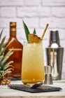Gelber Cocktail im Glas garniert mit Ananasstück und grünen Blättern mit Papierstroh auf Schieferuntersetzer mit Barlöffel gelegt — Stockfoto