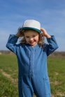 Menina feliz em roupas elegantes e boné olhando para a câmera enquanto está em pé na grama no dia ensolarado de verão no campo — Fotografia de Stock