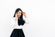 Adolescente étnica asiática mujer en traje formal de pie cerca de la pared blanca hablando en el teléfono móvil - foto de stock